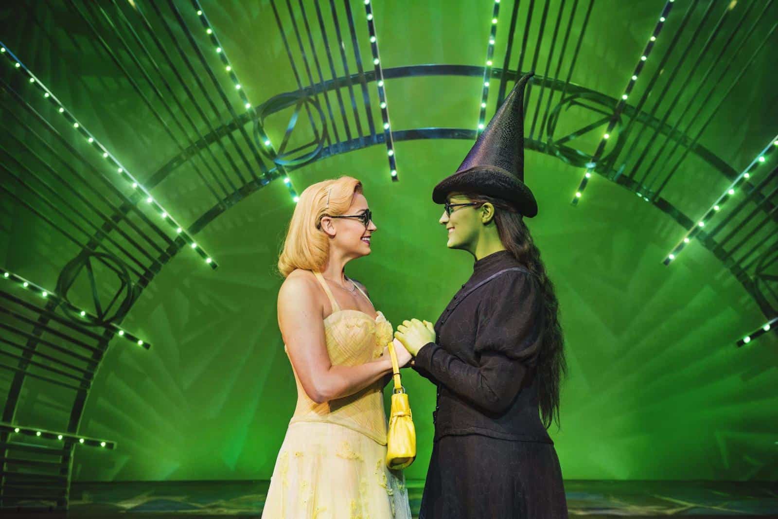 Glinda and Elphaba holding hands, smiling, and facing each other in the Emerald City