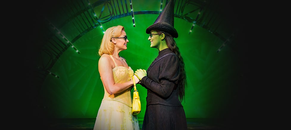Glinda and Elphaba in the Emerald City facing each other, holding hands, and smiling