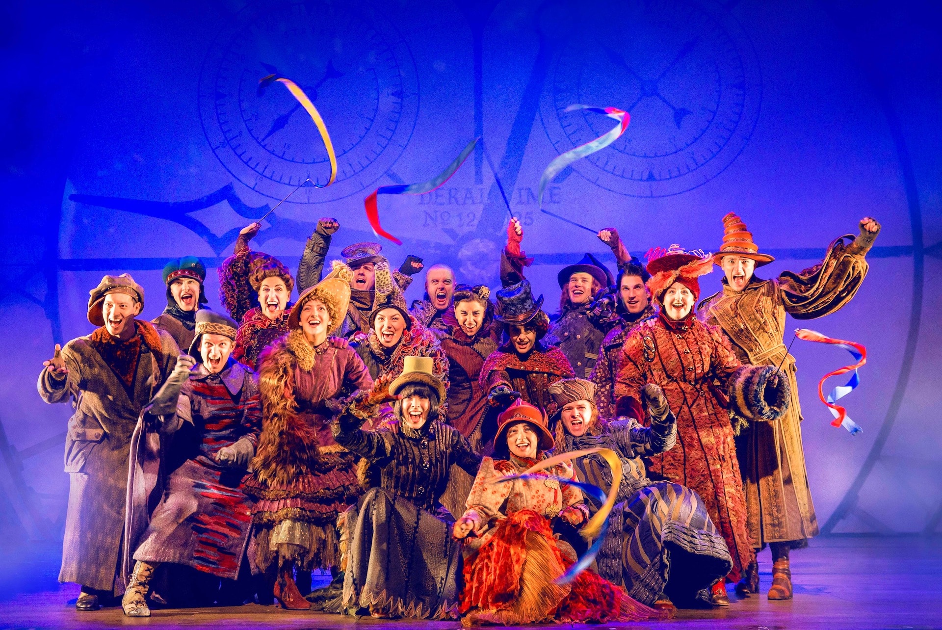 A Wicked London production image showing a group of Ozians waving ribbons and smiling on stage