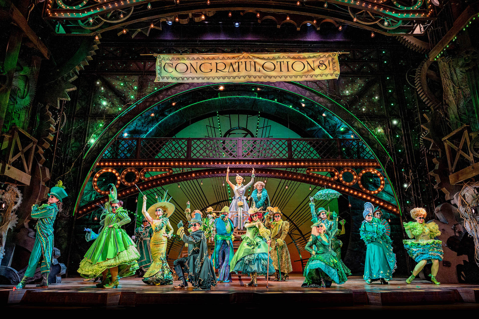 Wicked London production image showing The Emerald City Ozians listening to Glinda's speech with a banner that says "Congratulotions"