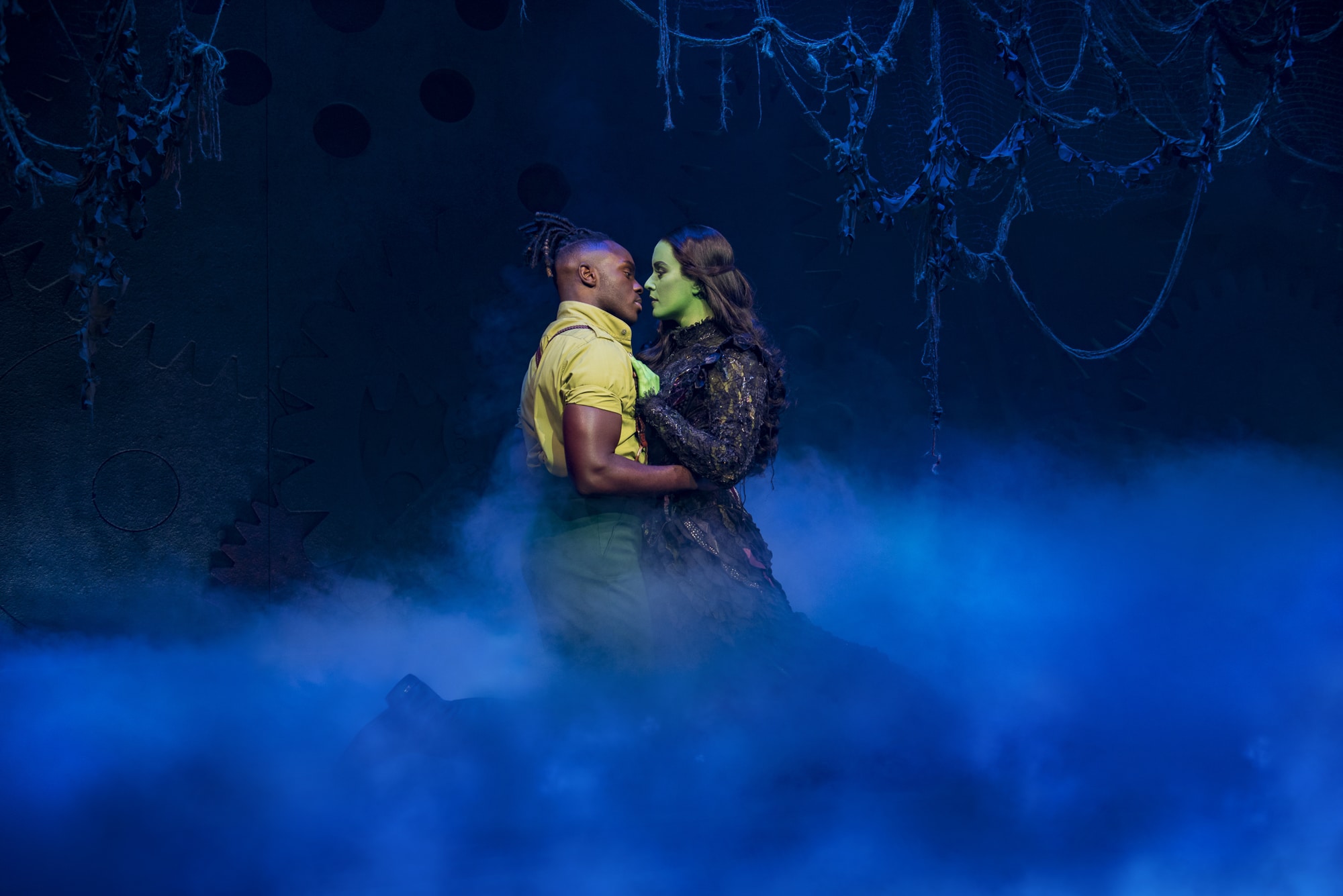 A Wicked London production image showing Fiyero and Elphaba embracing amongst vines, trees, and fog