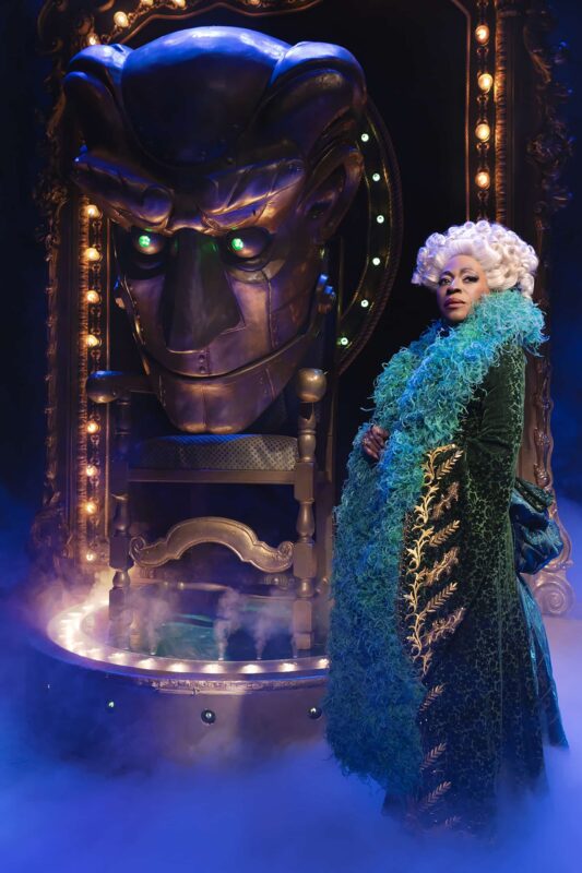 Madame Morrible in green coat with fluffy collar in front of Gold Oz head
