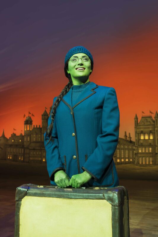 Elphaba smiling looking up holding suitcase in blue blazer