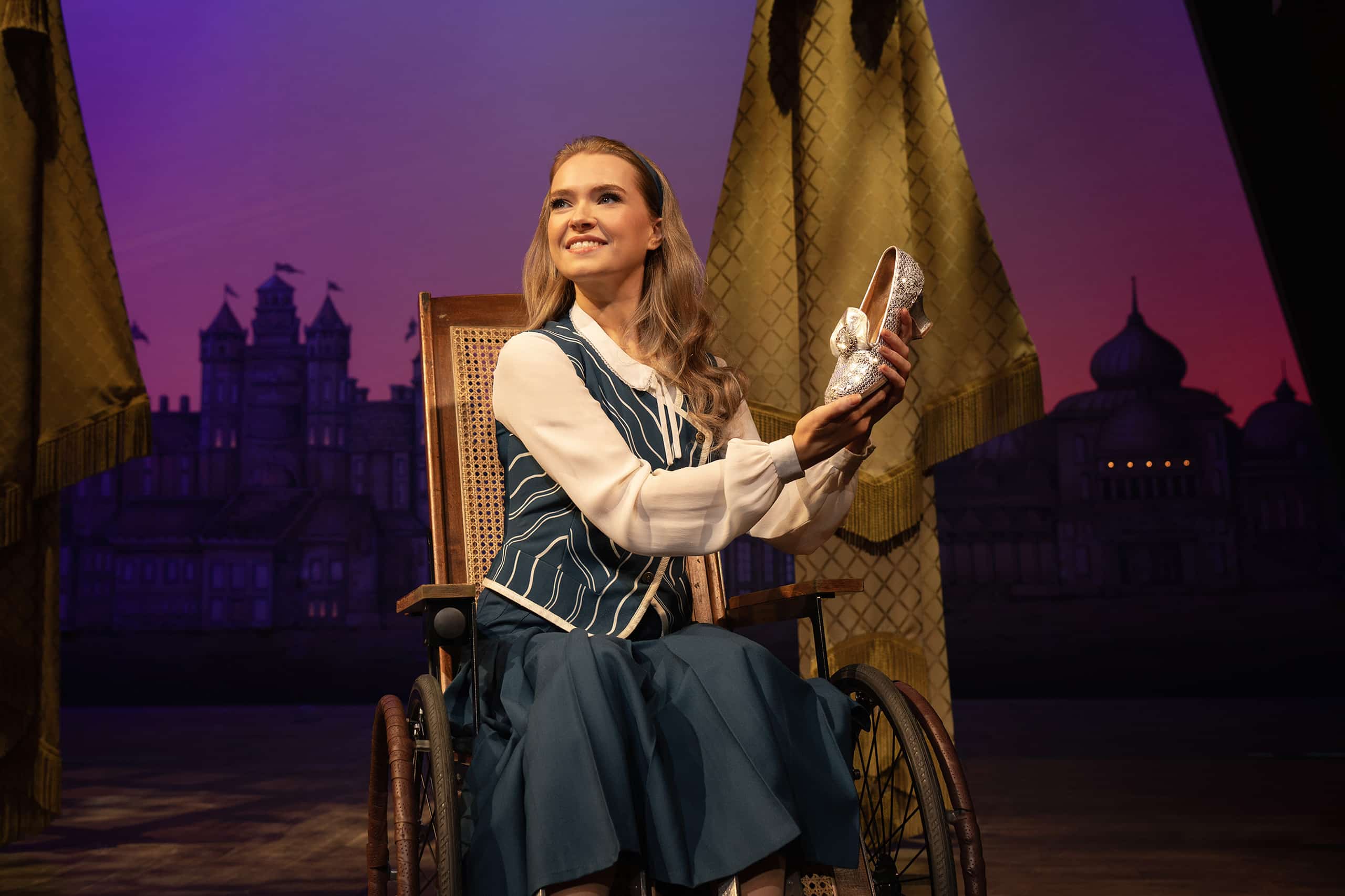 Nessarose in wheelchair holding silver sequinned shoes, smiling wearing blue striped top and skirt