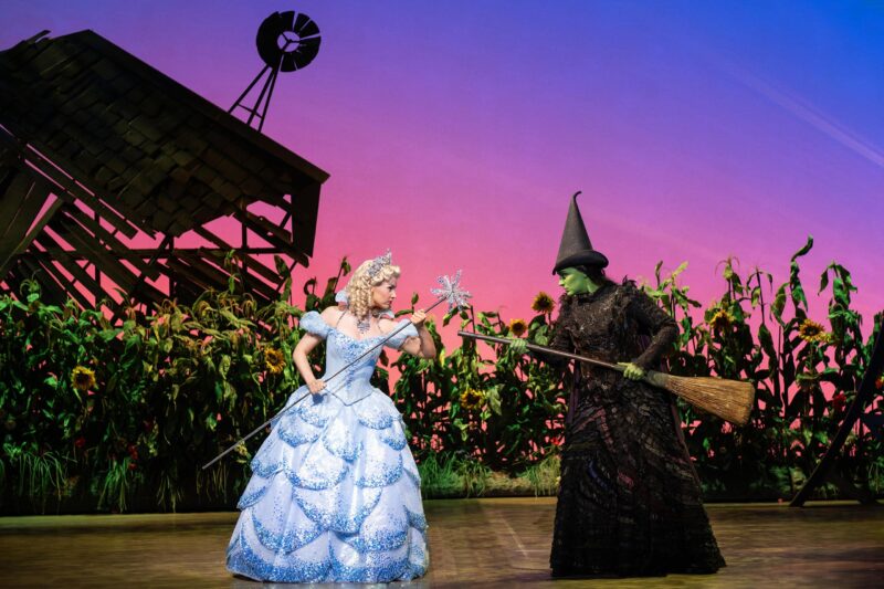 Glinda in blue ballgown pointing wand at Elphaba. Elphaba pointing broom at Glinda wearing black witch hat and black dress