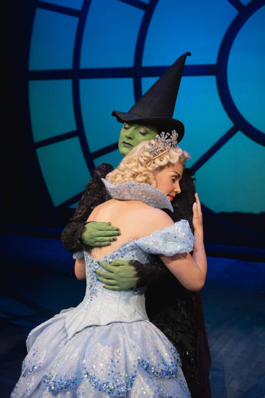 Elphaba wearing black dress and black witch hat hugging Glinda in blue ballgown with eyes shut in front of blue circular window