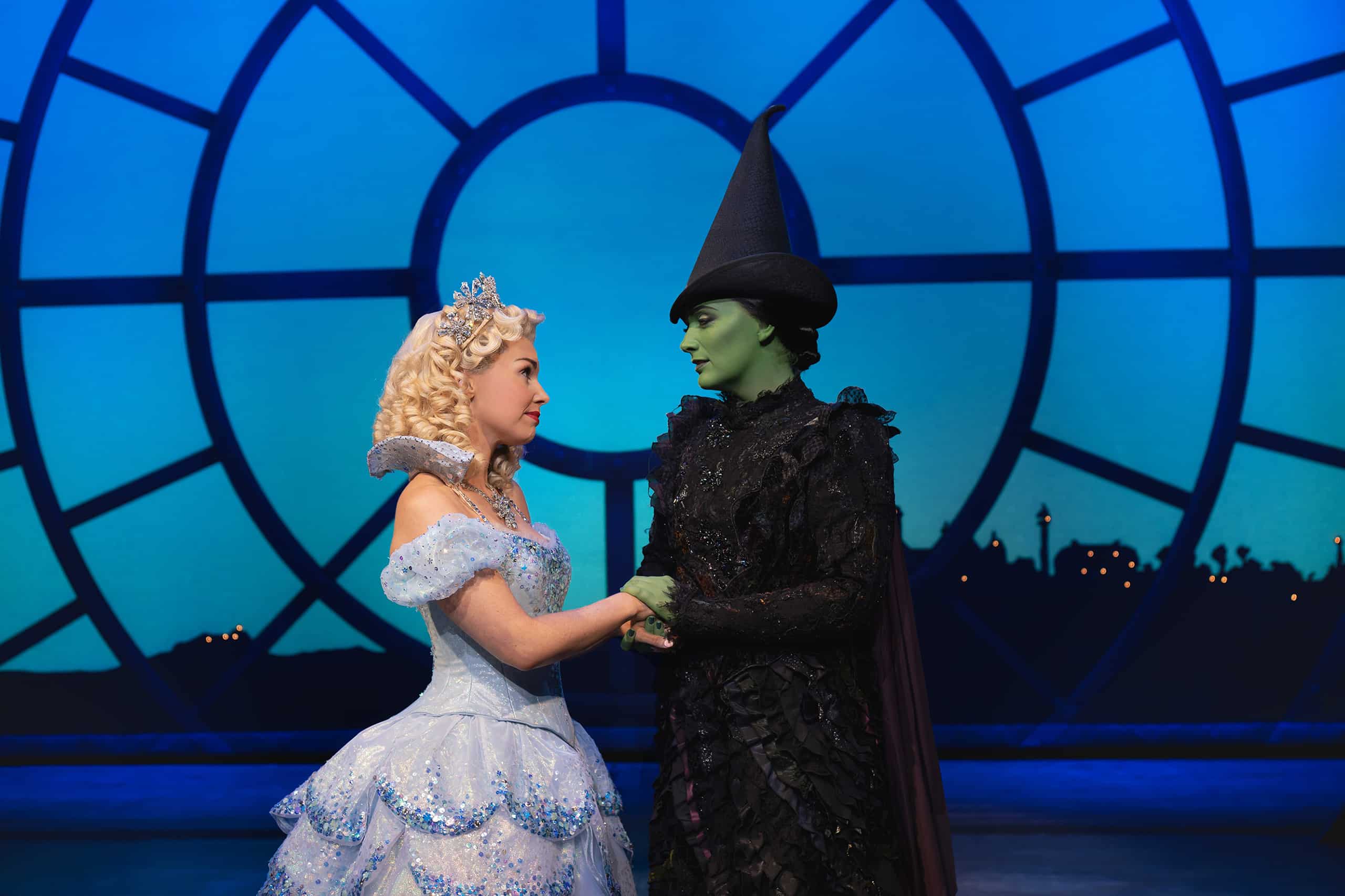 Glinda in blue ball gown holding hands and looking at Elphaba in black witch's hat and black dress