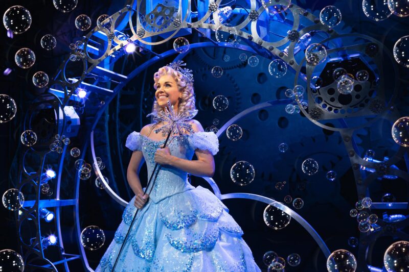 Glinda in bubble wearing blue ballgown holding wand and smiling