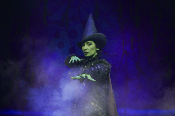 Green skinned girl wearing black dress, cape and witches hat gesturing towards the camera as if casting a spell.