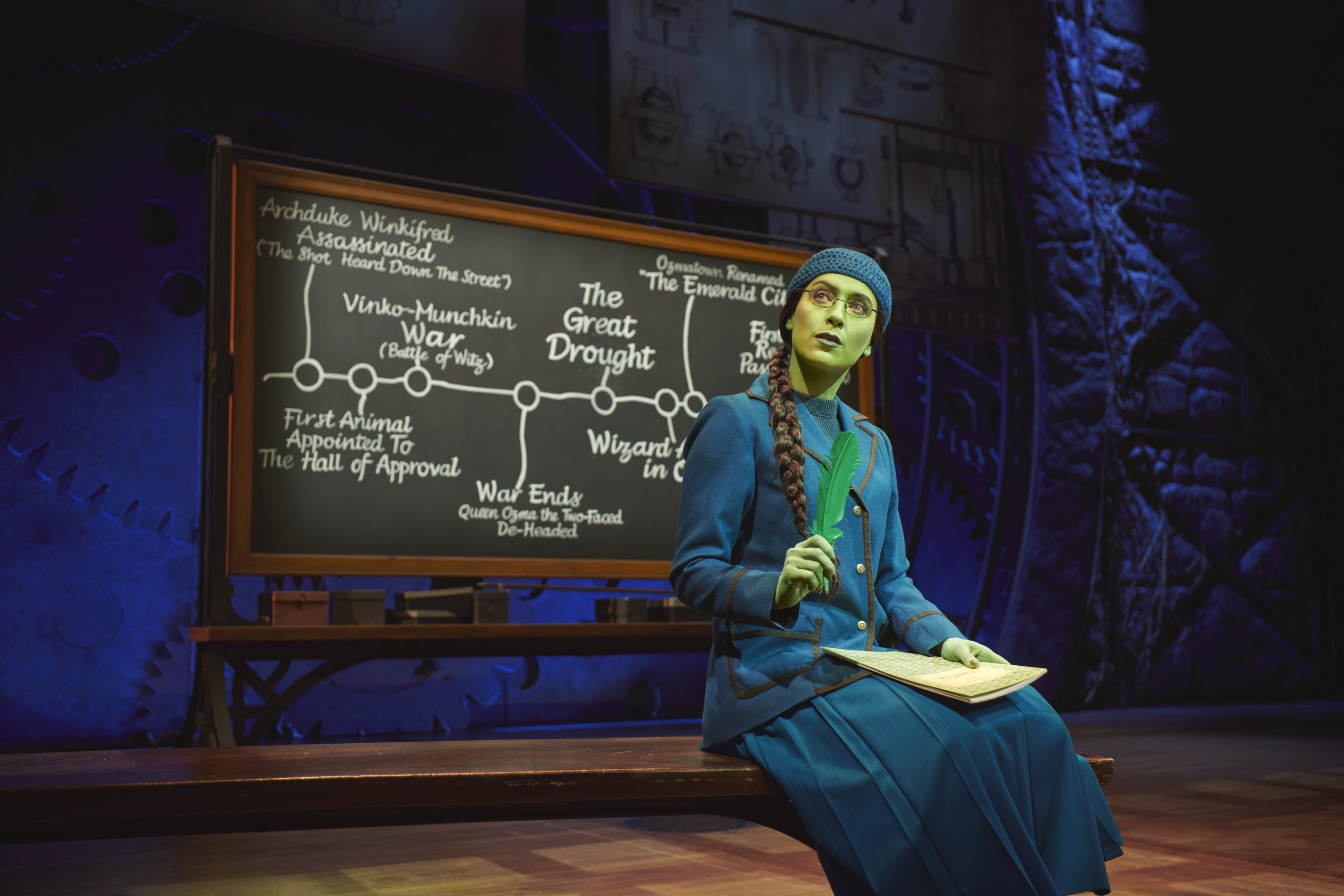 Laura Emmitt as a Young Elphaba in classroom with chalkboard behind, holding a green quill, she wears a navy crochet hat and navy blazer and long pleated skirt, her hair is plaited to the side and she has green skin.