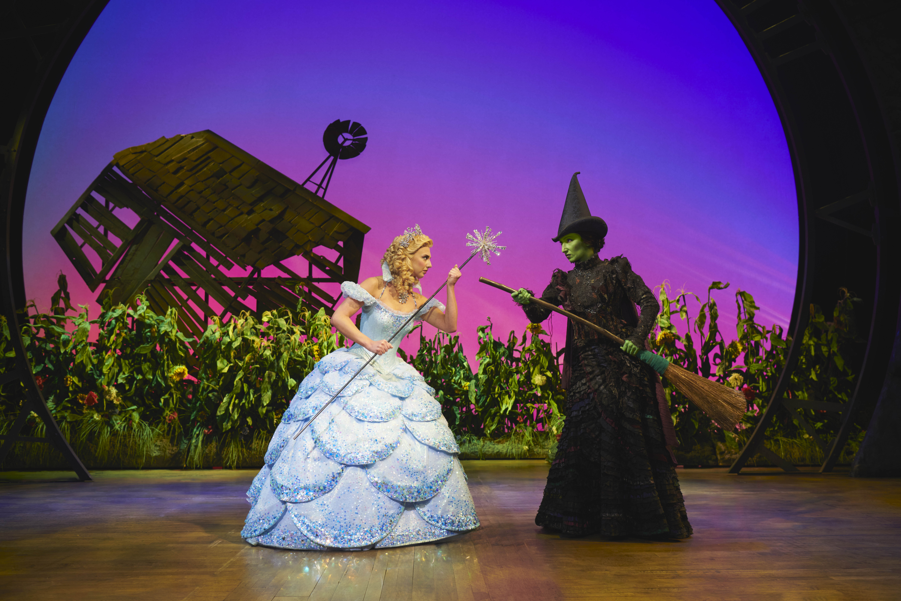Glinda with wand and Elphaba with broom facing one another, ready to fight. A field of sunflowers in the background and a fallen wooden house.