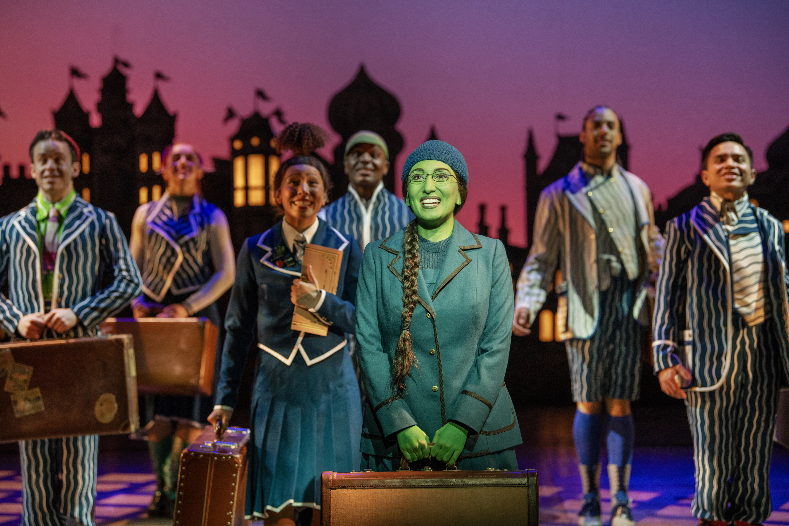 AN image of Emma Kingston as Elphaba in the musical Wicked holding a suitcase and surrounded by members of the company