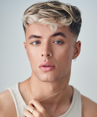 A young person with short, wavy, blond-highlighted hair and blue eyes looks at the camera. They wear a light tank top, earrings, and a ring, holding a thin necklace near their neck. The background is neutral and soft-focused.