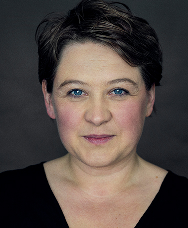 A person with short brown hair and blue eyes looks directly at the camera, smiling softly. They are wearing a black top and are posed against a dark, plain background.