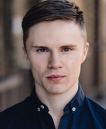A young man with short brown hair and fair skin stares directly at the camera. He wears a dark buttoned shirt and stands in front of a blurred outdoor background.