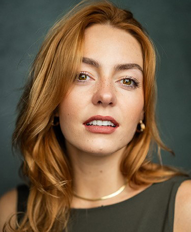 A woman with long auburn hair and light skin looks at the camera, wearing a dark top, gold hoop earrings, and a gold necklace, with a soft green background.