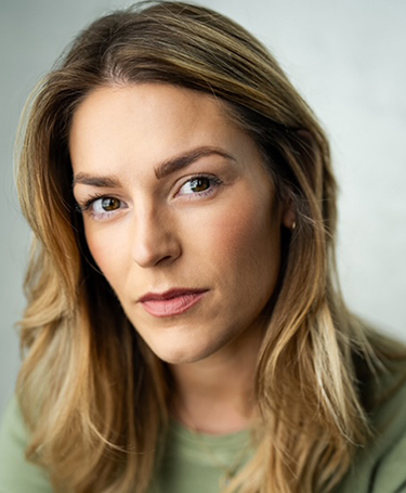 A woman with long, light brown hair and wearing a green top looks directly at the camera with a neutral expression against a softly blurred background.