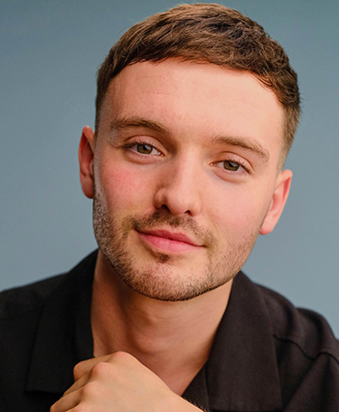 A young man with short brown hair and light facial hair looks directly at the camera, wearing a black shirt against a plain blue background. He has a relaxed expression and is resting his chin on his hand.