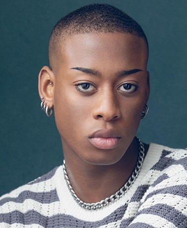 A young person with closely cropped hair, wearing hoop earrings, a chunky chain necklace, and a striped sweater, looks directly at the camera against a plain, dark background.
