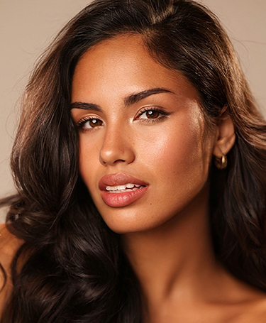 A woman with long, wavy dark hair and glowing skin looks at the camera with a gentle expression. She wears subtle makeup and small gold hoop earrings against a neutral background.