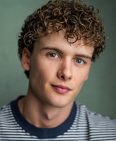 A young man with curly brown hair and blue eyes looks at the camera. He is wearing a striped shirt and is posed against a plain, muted green background.