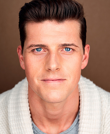 A man with short brown hair and blue eyes is looking directly at the camera. He is wearing a white knit sweater over a grey shirt, and the background is a plain, warm brown color.
