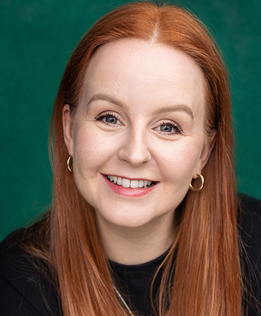 A woman with long straight red hair, fair skin, and blue eyes smiles warmly at the camera. She is wearing gold hoop earrings and a black top, with a green background behind her.