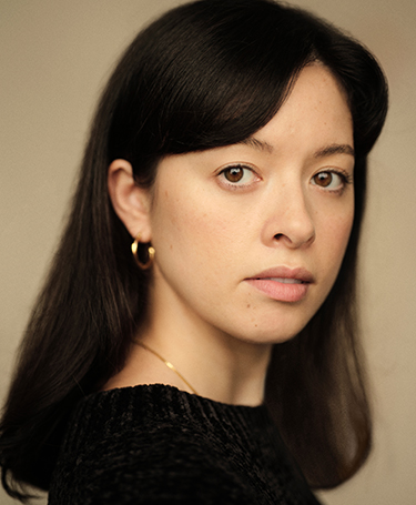 A woman with long dark hair, wearing gold hoop earrings and a black top, looks at the camera with a neutral expression against a beige background.