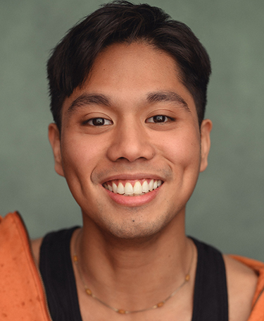 A young person with short dark hair, brown eyes, and a wide smile showing dimples, wearing an orange jacket over a black shirt and a beaded necklace, posed in front of a plain green background.