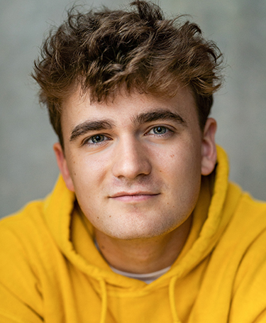 A young man with wavy brown hair and blue eyes, wearing a yellow hoodie, looks directly at the camera with a neutral expression. The background is softly blurred.