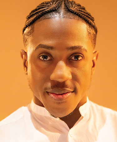 A person with neatly braided hair, wearing a white collared shirt, smiles softly at the camera against a warm, orange background.