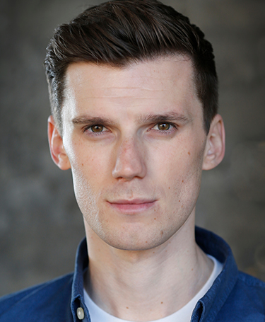 A man with short brown hair and fair skin is looking directly at the camera with a neutral expression. He is wearing a blue collared shirt over a white t-shirt, and the background is blurred and gray.