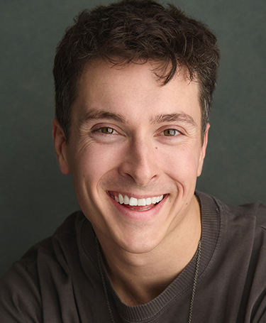 A young man with short, curly brown hair and light skin smiles warmly at the camera. He is wearing a dark-colored crew neck shirt and is posed against a plain, dark background.