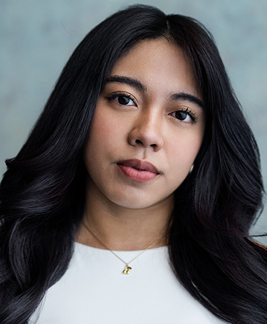A woman with long, wavy black hair wearing a white top and a gold necklace looks at the camera with a neutral expression, against a soft, blurred light blue background.