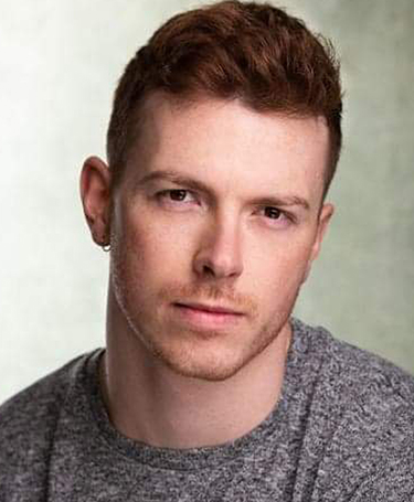 A young man with short reddish-brown hair and light skin, wearing a gray shirt, looks directly at the camera with a neutral expression against a softly blurred background.